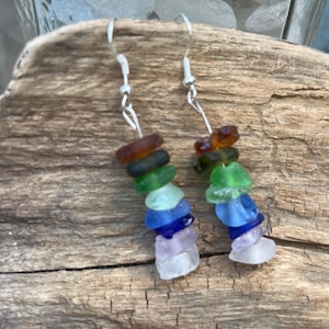 May include: Handmade earrings featuring colorful sea glass pieces in shades of brown, green, blue, and purple. The sea glass is strung on silver-colored hooks, creating a unique and eye-catching accessory. The earrings are displayed on a piece of driftwood.