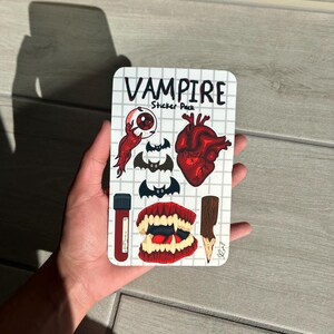 May include: A sticker pack with a vampire theme. The pack includes stickers of a bloody eyeball, bats, a heart, a vial of blood, vampire teeth, and a stake. The words "Vampire Sticker Pack" are printed on the top.
