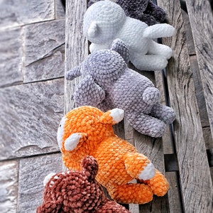 May include: Five crocheted stuffed animals in different colors, including brown, orange, gray, white, and purple, are lined up on a wooden bench. The animals are all facing the same direction and have a similar design.