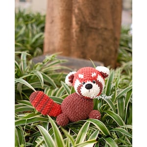 Op de afbeelding: Een gehaakt rood panda pluche speelgoed met een wit gezicht en zwarte ogen. Het speelgoed zit in een bed van groen-wit gestreepte bladeren.