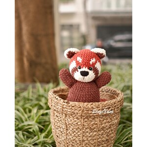 Op de afbeelding: Een gehaakt rood panda pluche speelgoed dat in een geweven mand zit. De panda is bruin met een rode buik en een wit gezicht.