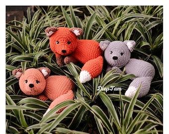 May include: Three crocheted fox plushies in different colors: orange, pink, and gray. The plushies are lying on a bed of green leaves. The plushies have white yarn for their tails and noses.