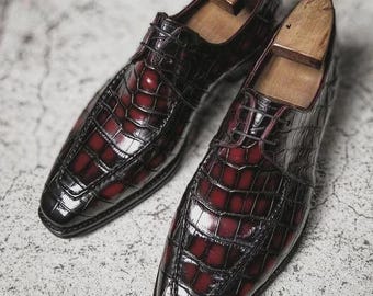 May include: A pair of dark burgundy leather dress shoes with a crocodile skin pattern. The shoes have a glossy finish and are laced up. Wooden shoe trees are inserted inside the shoes.