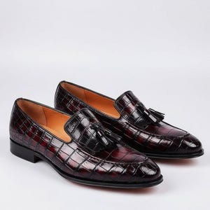 May include: A pair of burgundy leather loafers with a crocodile skin texture. The slip-on shoes have a tassel detail and a low heel. The interior is a light brown color.