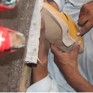 K&ouml;nnte beinhalten: Nahaufnahme der H&auml;nde eines Schuhmachers, der an einem gelben Lederschuh arbeitet. Der Schuh ist teilweise montiert, wobei die Sohle und der obere Teil zusammengen&auml;ht werden. Der Hintergrund ist unscharf, was die Handwerkskunst hervorhebt.