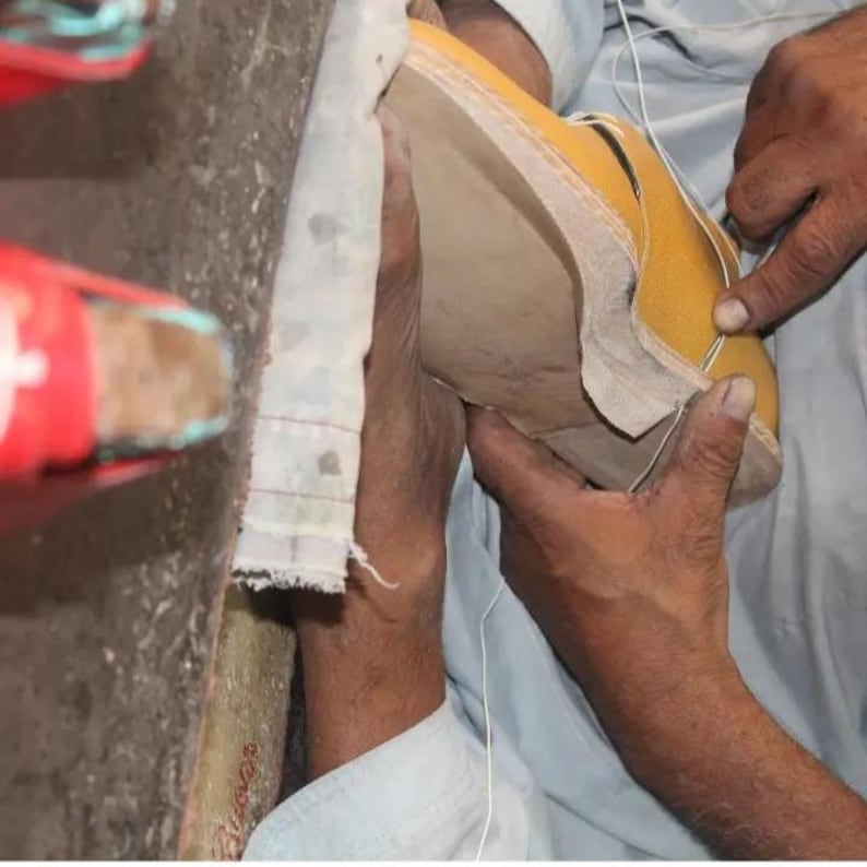 K&ouml;nnte beinhalten: Nahaufnahme der H&auml;nde eines Schuhmachers, der an einem gelben Lederschuh arbeitet. Der Schuh ist teilweise montiert, wobei die Sohle und der obere Teil zusammengen&auml;ht werden. Der Hintergrund ist unscharf, was die Handwerkskunst hervorhebt.