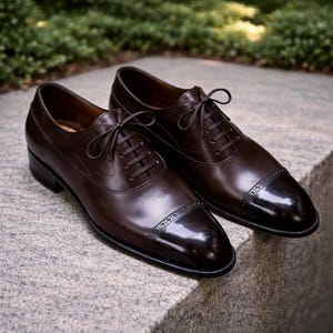 May include: A pair of polished brown leather dress shoes with a cap toe design. The shoes have a lace-up closure and are displayed on a stone surface. The shoes are a classic style, suitable for formal occasions.