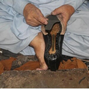 K&ouml;nnte beinhalten: Nahaufnahme einer Person, die an einem schwarzen Lederschuh arbeitet. Der Schuh wird mit einer dunklen Sohle und einem hellfarbigen Innenraum zusammengebaut. Die Person tr&auml;gt ein hellblaues Hemd.