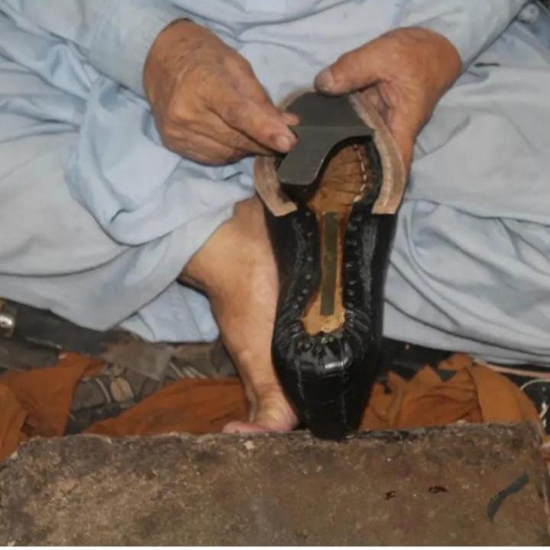 K&ouml;nnte beinhalten: Nahaufnahme einer Person, die an einem schwarzen Lederschuh arbeitet. Der Schuh wird mit einer dunklen Sohle und einem hellfarbigen Innenraum zusammengebaut. Die Person tr&auml;gt ein hellblaues Hemd.