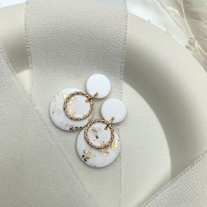 Könnte beinhalten: Weiße und goldfarbene Ohrringe mit einem geschichteten Design. Jeder Ohrring hat eine große, runde, weiße Basis mit Goldblatt-Akzenten, einen goldenen Ring und einen kleineren weißen Kreis. Die Ohrringe sind mit goldfarbener Hardware verbunden.
