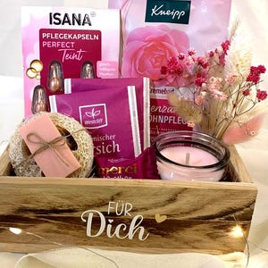Könnte beinhalten: Eine hölzerne Geschenkbox mit der Aufschrift "FÜR Dich", gefüllt mit Isana Perfect Teint Kapseln, einem Kneipp Rosenbad, Westcliff Tee, einem Merci Schokoriegel, einer rosa Kerze im Glas, einem Luffa mit rosa Seifenstück und getrockneten Blumen.