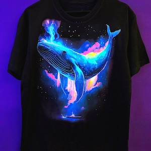 Puede incluir: Camiseta negra con una vibrante ilustración de una ballena en un entorno cósmico. La ballena está representada en tonos azules, con nubes coloridas y estrellas. Un pequeño barco es visible debajo, agregando profundidad al diseño.