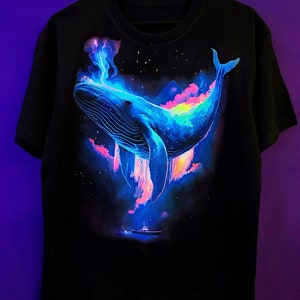 May include: Black t-shirt with a glow-in-the-dark design of a blue whale swimming in a starry sky with pink and purple clouds.