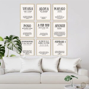 May include: A living room scene featuring a white sofa, armchair, and small side table. Nine framed prints with black text hang above the sofa. The prints feature Hawaiian words and phrases. Large green plants add a natural touch to the room.