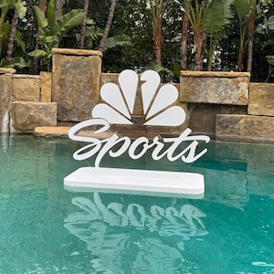 May include: A white floating sign in a pool with the word "Sports" in cursive script. The sign features a stylized white peacock logo. The pool water is a clear turquoise color, and the background includes stone walls and lush greenery.