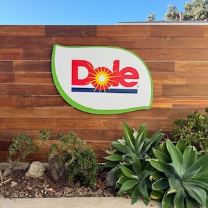 May include: A large sign with the Dole logo, featuring the word "Dole" in red letters with a yellow sun graphic, is mounted on a wooden fence. The sign is leaf-shaped with a green border. Green plants and shrubs are in front of the fence.