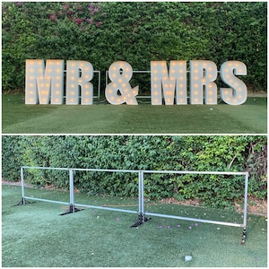 May include: White light-up letters spelling "MR & MRS" on a green lawn. The letters are illuminated with warm white lights. A metal fence is in the foreground.