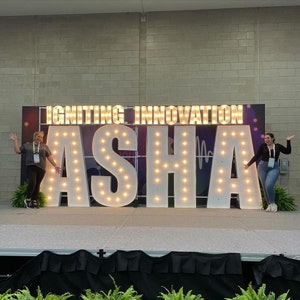 May include: Two people stand in front of a large illuminated sign that reads "IGNITING INNOVATION ASHA". The sign is made of white letters with light bulbs outlining the letters. The people are smiling and appear to be happy.