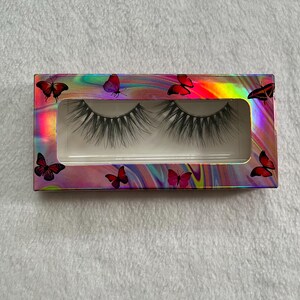 May include: A pair of false eyelashes presented in a rectangular box. The box has a holographic, iridescent finish with red butterfly accents. The eyelashes are black and appear full and voluminous.