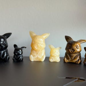 May include: Five carved figurines of a cartoon character, each in a different color. The figurines are arranged in a row from left to right: black, yellow, brown, yellow, and brown. The figurines are all in the same pose, with their arms outstretched and their mouths open in a smile.