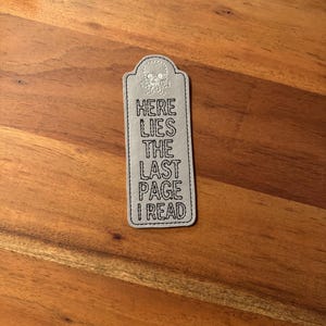 May include: A gray leather bookmark with a skull design and the text "Here Lies The Last Page I Read".