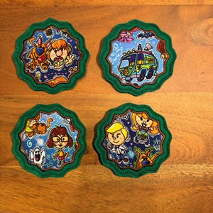 May include: Four round coasters with a green outer edge and a brown inner edge. Each coaster features a different cartoon image from the Scooby Doo franchise. One coaster features Scooby Doo and Shaggy, another features the Mystery Machine, another features Velma and Scooby Doo, and the last features Daphne and Fred.