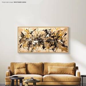 Puede incluir: Pintura abstracta en tonos negros, blancos y dorados. La pintura está enmarcada en un marco de madera natural y cuelga de una pared blanca sobre un sofá marrón.