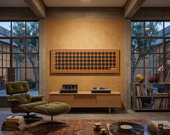 Op de afbeelding: Een rechthoekig wandkunstwerk met een raster van zwarte cirkels op een oranje achtergrond, met de verticale tekst "bauhaus" aan de linkerkant en "1919" aan de rechterkant, tentoongesteld in een kamer met een houten mediameubel en een olijfgroene fauteuil.