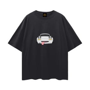 May include: Black t-shirt featuring a cartoon car with a white body, black accents, and a pink tongue sticking out. The car has headlights and a smiling expression. The shirt has a crew neck and short sleeves.