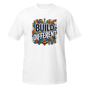 Peut inclure: T-shirt blanc avec un motif graphique coloré « BUILD DIFFERENT ». Le design présente un thème de construction avec des engrenages, des blocs et une grue dans des tons de bleu, orange, jaune et vert.