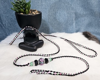 May include: A black and white braided cord with a pink and green floral bead. The cord is looped and has a small loop at one end.