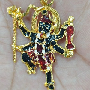May include: A gold pendant depicting a deity with a black face and limbs, red and white details, and a red headdress. The pendant has intricate detailing and hangs from a fine gold chain. The figure is holding objects.