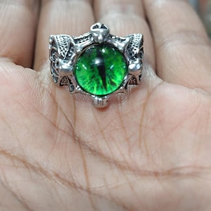 May include: A silver ring with a green eye design. The ring has a detailed design with a skull and crossbones motif.