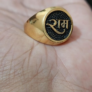 May include: A gold-colored ring with a black circular face. The face of the ring has the word "राम" in a stylized font. The ring is set on a light-colored background.