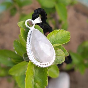 Può includere: Un ciondolo d'argento con una pietra preziosa bianca a forma di lacrima. Il ciondolo è su un cordoncino nero ed è circondato da foglie verdi.