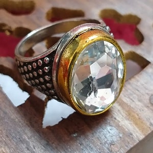 Op de afbeelding: Een zilveren ring met een grote, heldere kristal in een gouden bezel. De ring heeft een getextureerde band met kleine zilveren stippen.