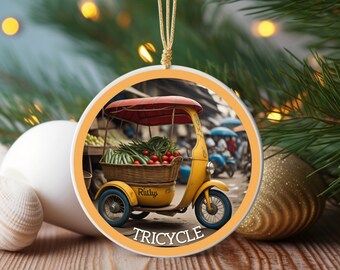 May include: Round Christmas ornament featuring a yellow tricycle with a red canopy, carrying a basket of vegetables. The word "TRICYCLE" is printed in white. The ornament is hanging with a gold string, with a blurred background of a Christmas tree and ornaments.