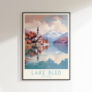 May include: A vintage travel poster showcasing a scenic view of Lake Bled in Slovenia. The poster depicts a picturesque island with a church and a reflection of the church in the water. The mountains in the background are snow-capped and the sky is a soft blue with fluffy clouds. The text "LAKE BLED" and "SLOVENIA" is printed in blue at the bottom of the poster.