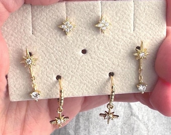 May include: A set of three gold star earrings with clear crystals. The earrings are displayed on a white card with a hole in the center. Two of the earrings are studs and one is a dangle earring with a small crystal at the bottom.