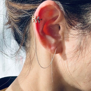 May include: A gold-toned ear cuff with a leaf design, adorned with small, clear gemstones. A delicate chain extends from the cuff, connecting to a straight bar earring. The jewelry is worn on the ear, with the chain and earring hanging down.