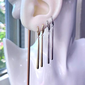 May include: Gold and silver-toned dangle earrings displayed on a white mannequin head. The earrings have a simple, elegant design with a long, slender bar hanging from a small hoop. The gold earrings are on the left, and the silver earrings are on the right.