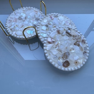 May include: A set of four round coasters with a mosaic design made of white and brown shell pieces. The coasters are in a gold-coloured metal stand with a curved handle. The coasters have a white border.