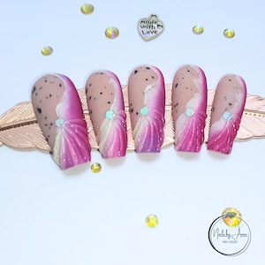 Könnte beinhalten: Ein Satz von fünf künstlichen Nägeln mit hellbeigem Grund, übersät mit schwarzen Punkten. Jeder Nagel hat ein weißes und rosa Design mit einem Schmuckakzent. Die Nägel sind auf einem goldfarbenen Dekorationsteil angeordnet, mit verstreuten Strasssteinen.