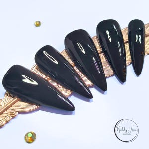 Könnte beinhalten: Ein Set aus vier glänzend schwarzen Press-on-Nägeln in spitzer Stiletto-Form, diagonal angeordnet auf einem strukturierten goldenen Display in Federform. Ein kleines kreisförmiges Logo in der rechten unteren Ecke zeigt Nails by Anne Nail Salon.