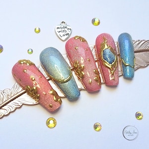 Könnte beinhalten: Ein Set aus sechs langen, ovalen künstlichen Nägeln mit einem pink- und blauglitzernden Finish. Die Nägel sind mit Blattgold und einem goldenen Akzent verziert. Die Nägel sind auf einem goldenen federförmigen Display angeordnet. Das Bild zeigt auch einen kleinen silbernen Anhänger mit dem Text "Made with Love".