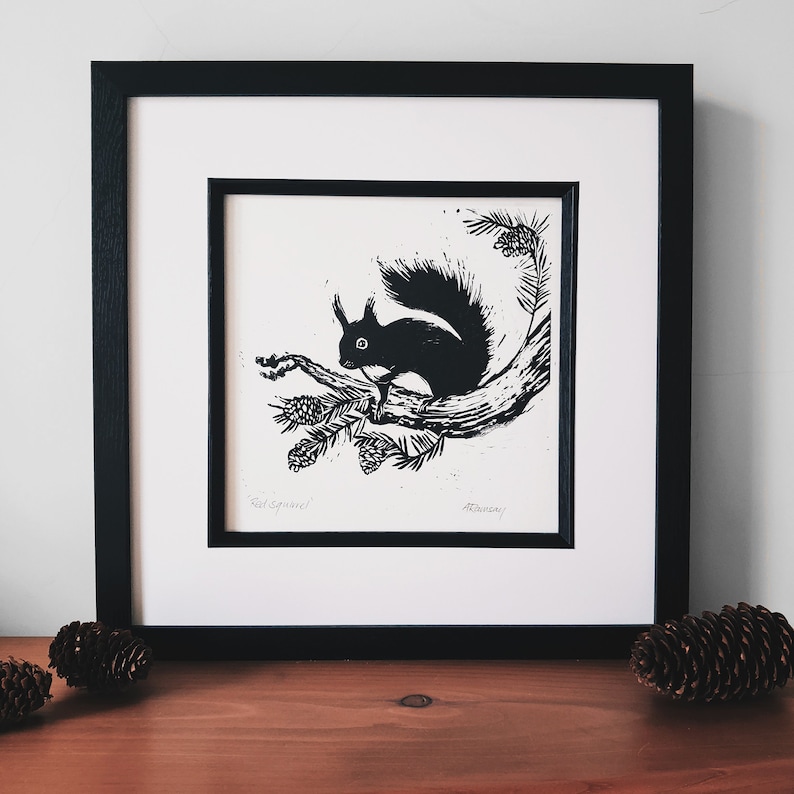 May include: Black and white print of a squirrel on a branch, framed in a black frame with a white mat. The print includes the text "Red Squirrel". The artwork is displayed on a wooden surface with pine cones.