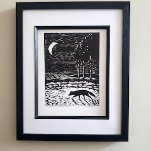 Peut inclure: Une estampe en linogravure en noir et blanc d'un renard marchant dans un champ sous un croissant de lune. L'estampe est encadrée dans un cadre noir avec un passe-partout blanc. Le titre de l'estampe est "The One The Shadows Fear" et le nom de l'artiste est "A Ramsay".