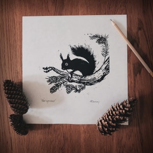 May include: Black and white print of a squirrel on a branch with pinecones and the text "Red Squirrel". A pencil and pinecones are on a wooden surface. The artwork is a linocut print.