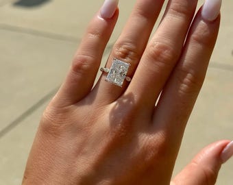Può includere: Un anello con diamante a taglio rettangolare con una grande pietra centrale e pietre laterali più piccole. L'anello è montato su una fascia sottile. La mano ha lo smalto bianco.
