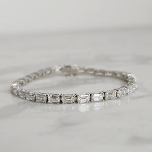 May include: A silver bracelet featuring a series of rectangular-cut, clear gemstones. The gemstones are set closely together, creating a continuous line of sparkle. The bracelet is designed with a secure clasp.